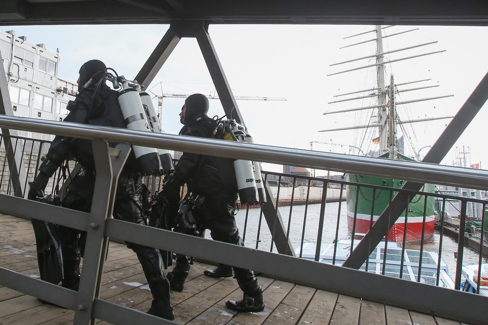 Taucher gehen an der Elbe in Hamburg über eine Brücke, im Hintergrund ist das Museumsschiff Rickmer Rickmers zu sehen.