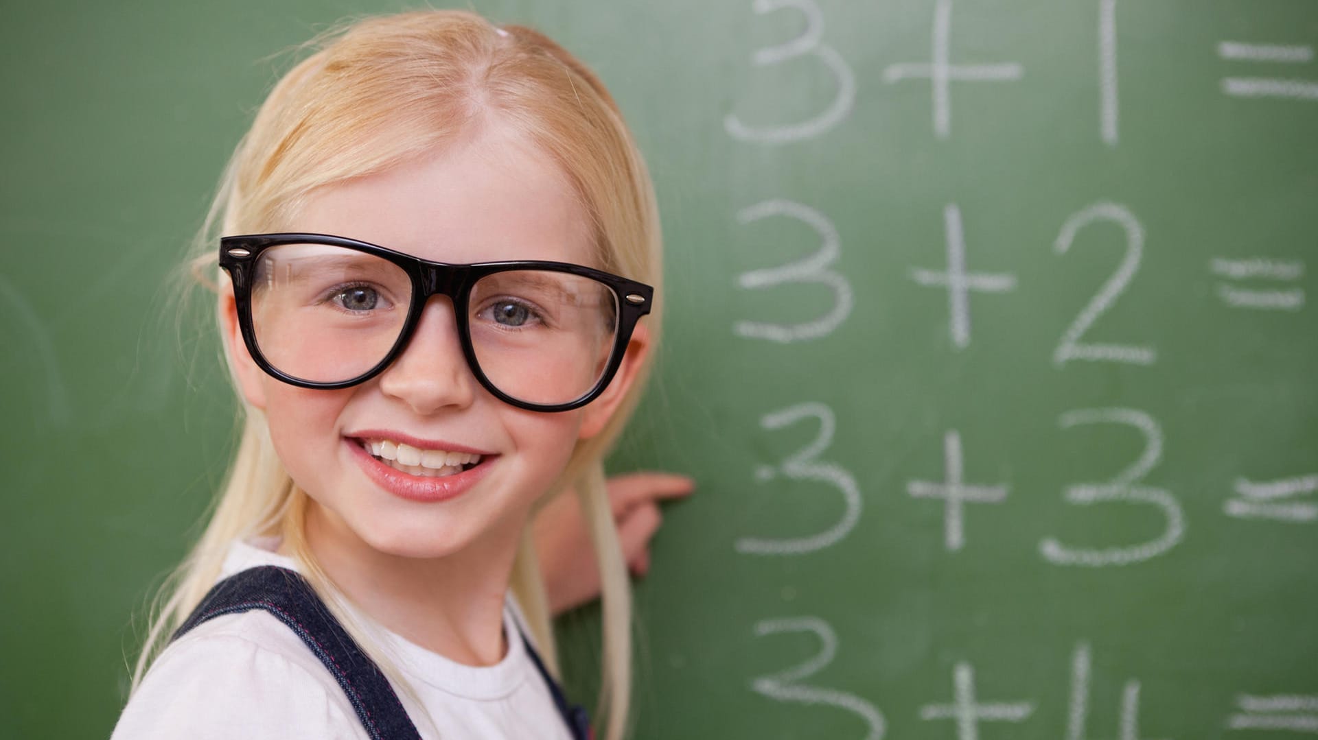 Ein Mädchen mit Hornbrille steht an einer Tafel.