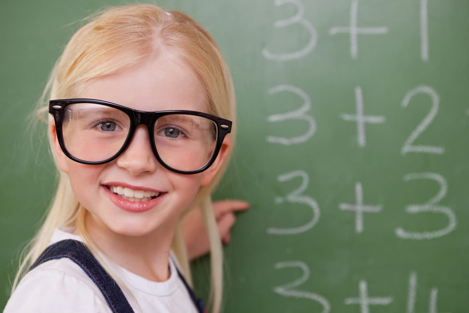 Ein Mädchen mit Hornbrille steht an einer Tafel.