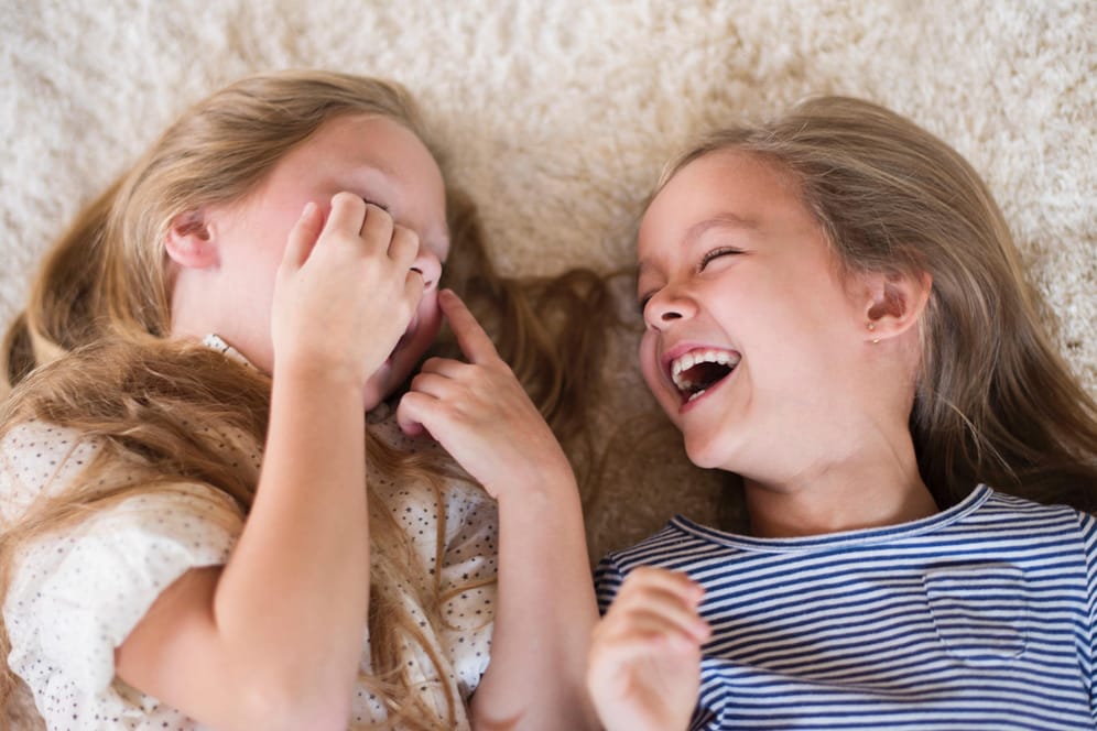 Zwei Mädchen liegen auf einem Teppich und lachen.