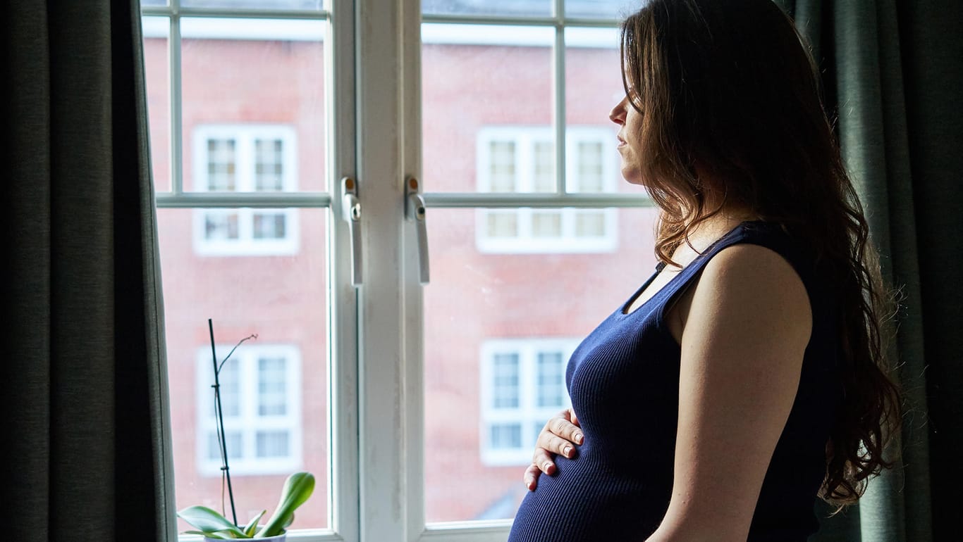 Eine Schwangere steht gedankenverloren an einem Fenster.
