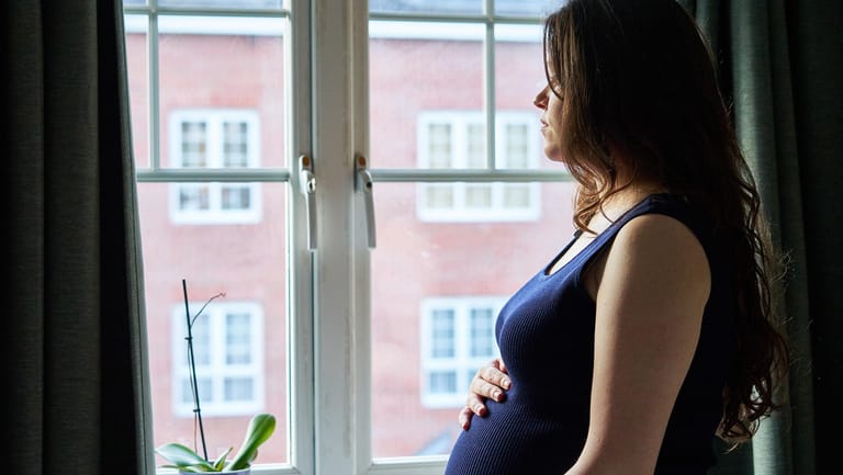 Eine Schwangere steht gedankenverloren an einem Fenster.