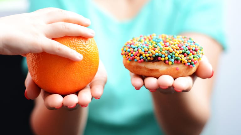 Eine Frau hält in der einen Hand eine Apfelsine, in der anderen einen Donut