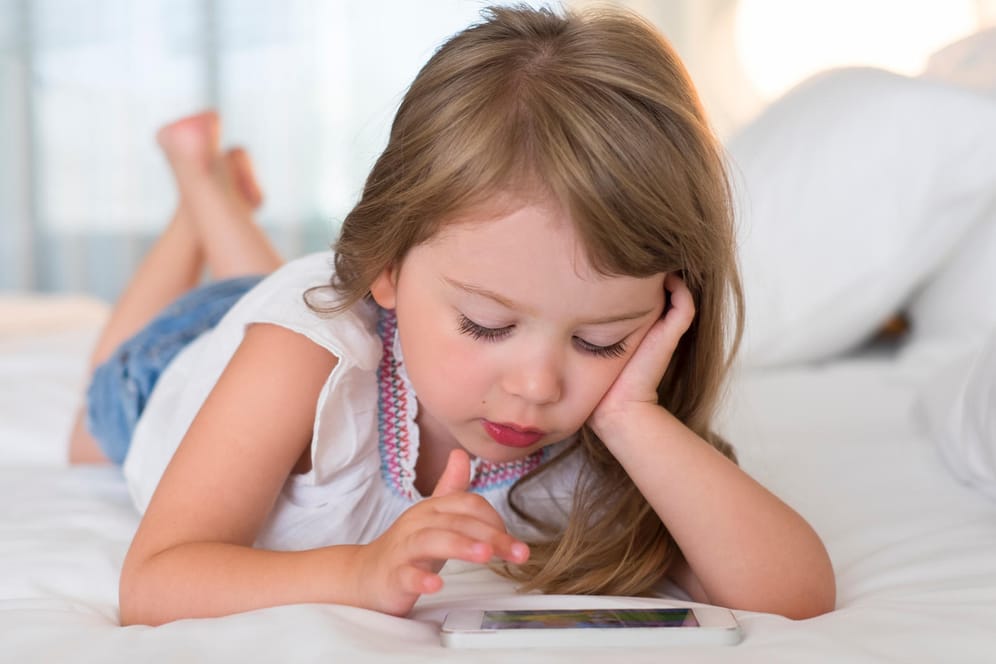 Ein Mädchen liegt auf dem Bett und schaut auf ein Smartphone.