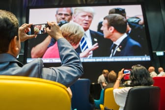Journalisten machen im Medienzentrum für Journalisten, das anlässlich des G20-Gipfels eingerichtet wurde, ein Foto von dem Aufeinandertreffen von Bundeskanzlerin Angela Merkel, dem französischen Staatspräsidenten Emmanuel Macron und US-Präsident Donald Trump, die live auf einen Bildschirm übertragen wird.
