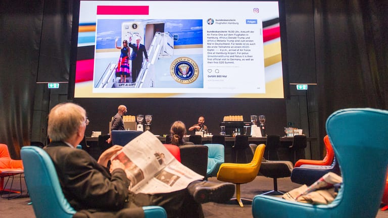 Eine Mann sitzt im Medienzentrum für Journalisten, das anlässlich des G20-Gipfels eingerichtet wurde, vor einem Bildschirm und liest eine Zeitung. Im Hintergrund ist die Ankunft von US-Präsident Trump und seiner Frau zu sehen.