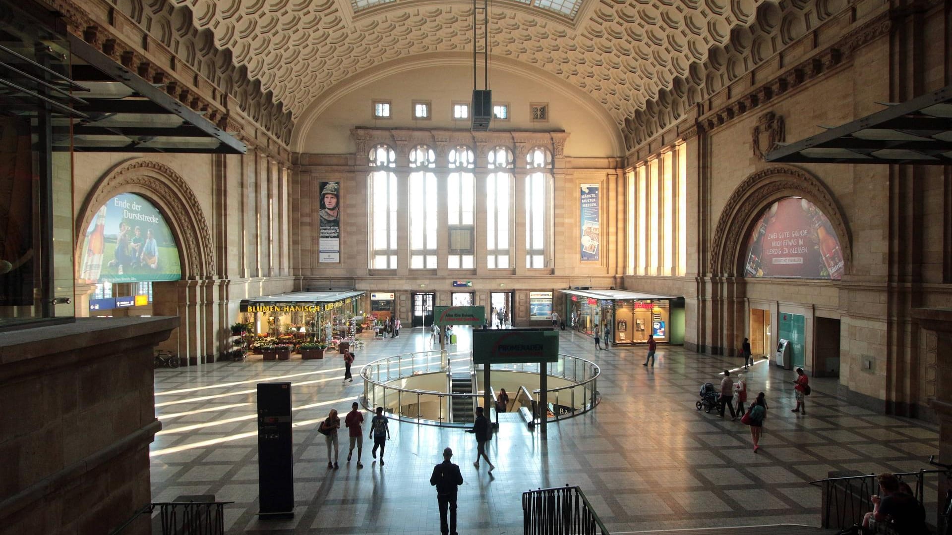 Fahrgäste und Passanten flanieren durch die Sonnenstrahlen durchflutete Westhalle des Leipziger Hauptbahnhofs.