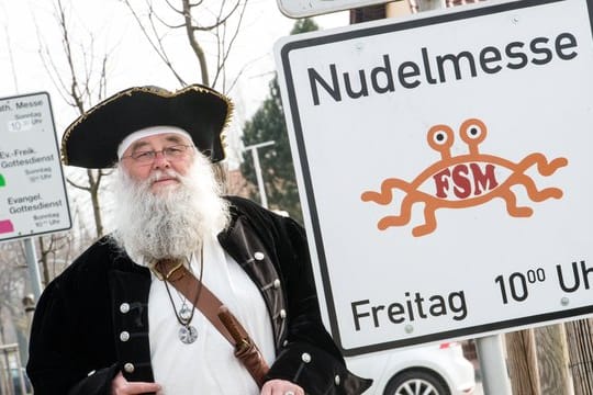 Rüdiger Weida vom Verein "Kirche der fliegenden Spaghettimonster" steht neben einem Schild, das für die "Nudelmesse" des Satirevereins im brandenburgischen Templin wirbt.