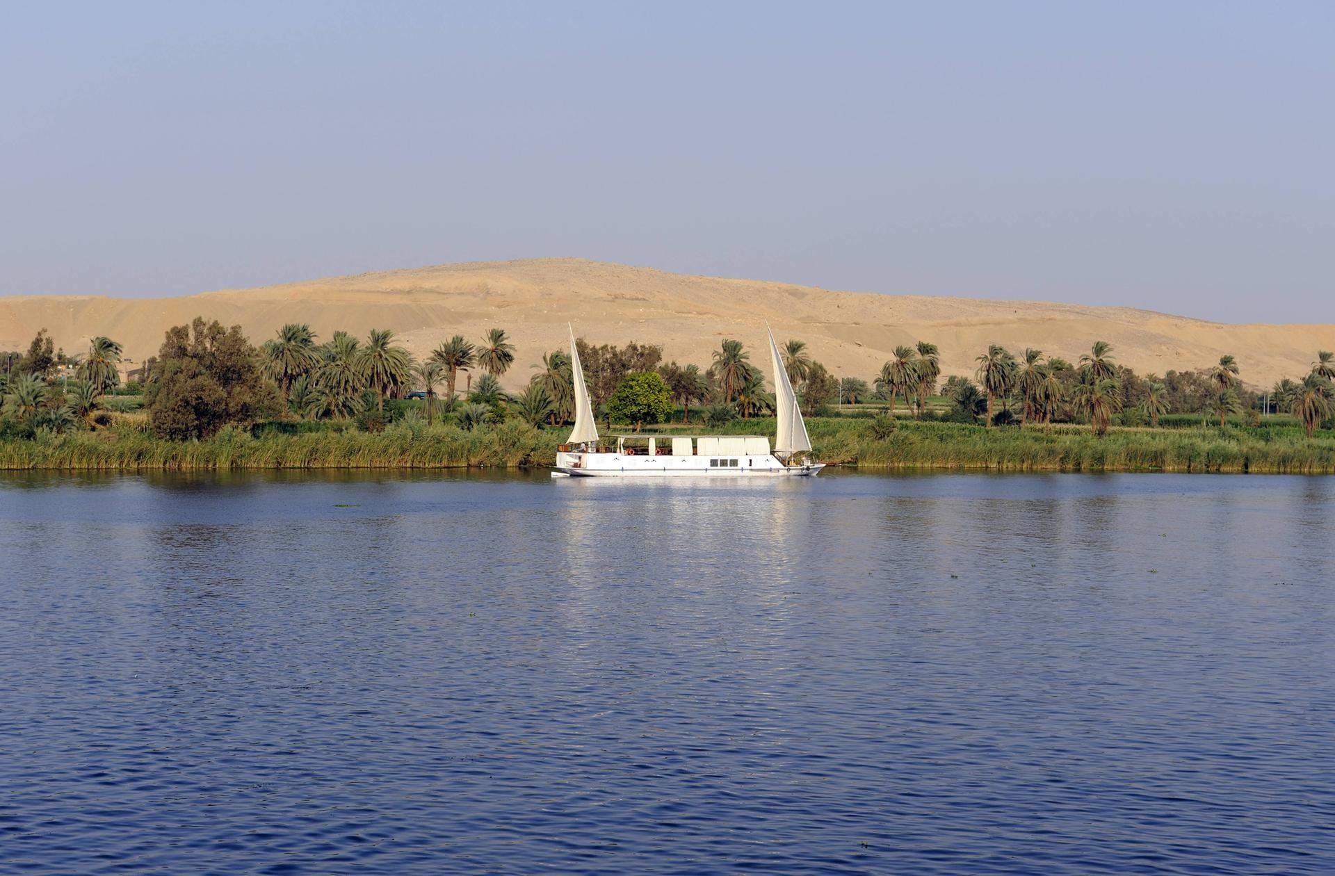 Zweimastige Felukke mit Dhau-Segeln auf dem Nil. Dahinter erstreckt sich die Sahara.