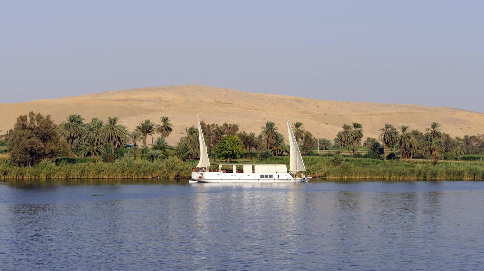 Zweimastige Felukke mit Dhau-Segeln auf dem Nil. Dahinter erstreckt sich die Sahara.