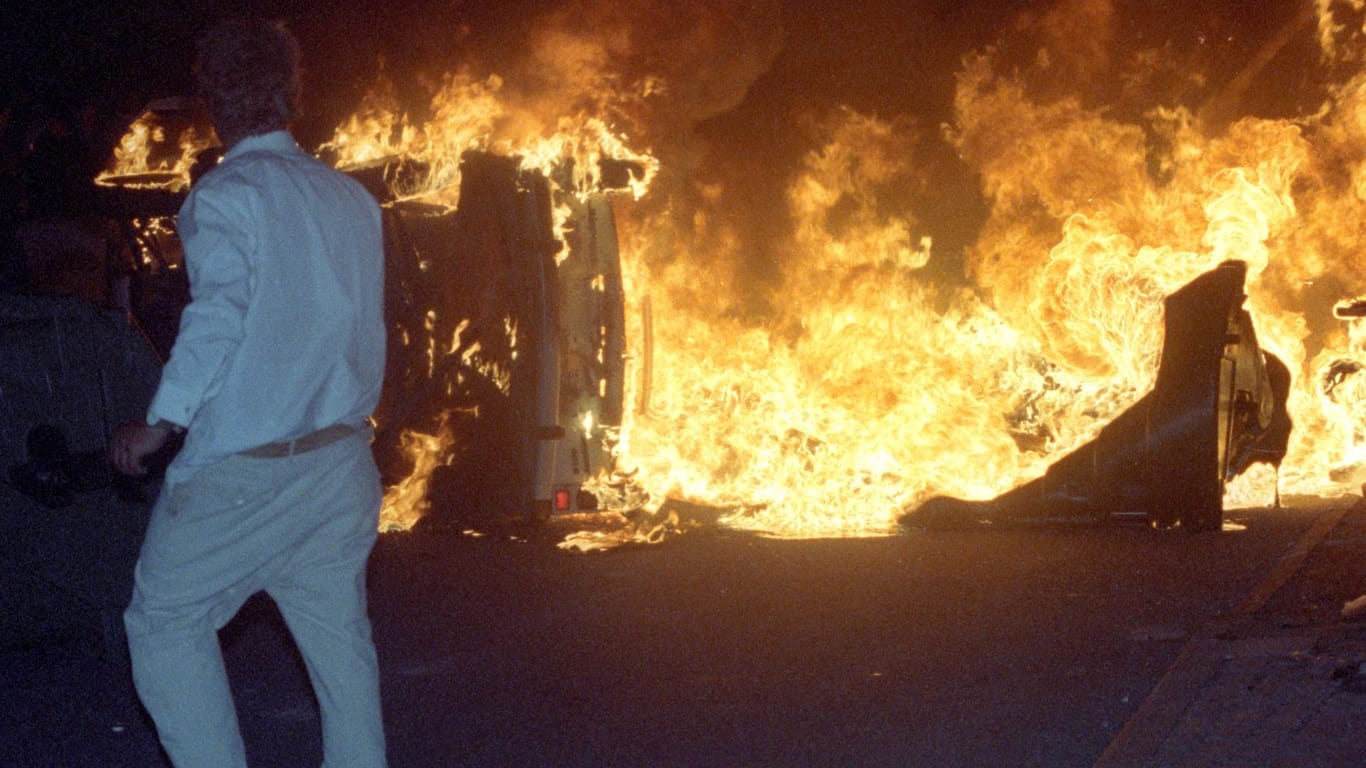 Ein Mann steht am 27.08.1992 vor einem brennenden Pkw auf einer Straße am zentralen Asylbewerberheim von Mecklenburg-Vorpommern in Rostock-Lichtenhagen.