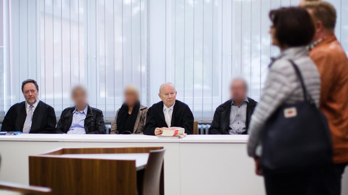 Die drei Angeklagten warten in Essen im Gerichtssaal neben ihren Anwälten auf den Prozessbeginn. Die Angehörigen des Opfers stehen rechts.