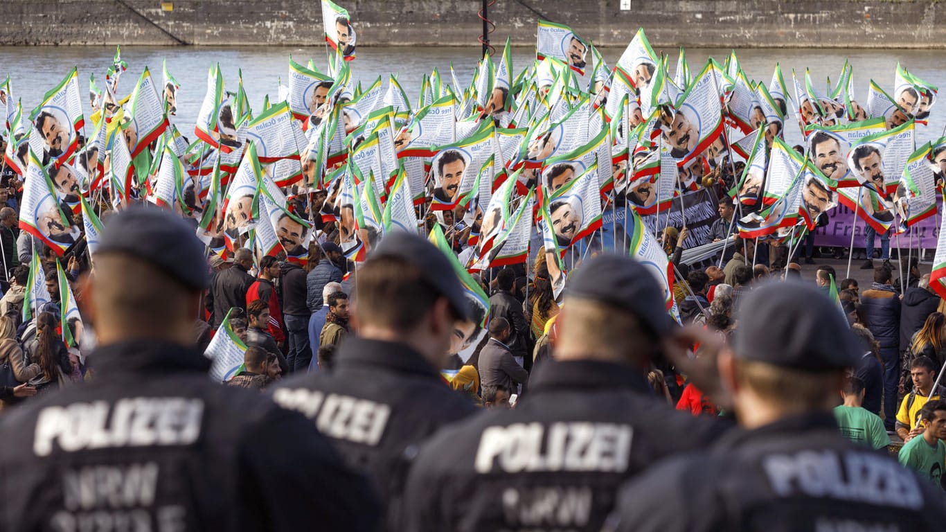 Beim Kurdischen Kulturfestivalt in Köln stehen Teilnehmer mit Fahnen, auf denen Porträts des PKK-Führers Öcalan zu sehen sind. Davor stehen Polizisten und beobachten die Situation.
