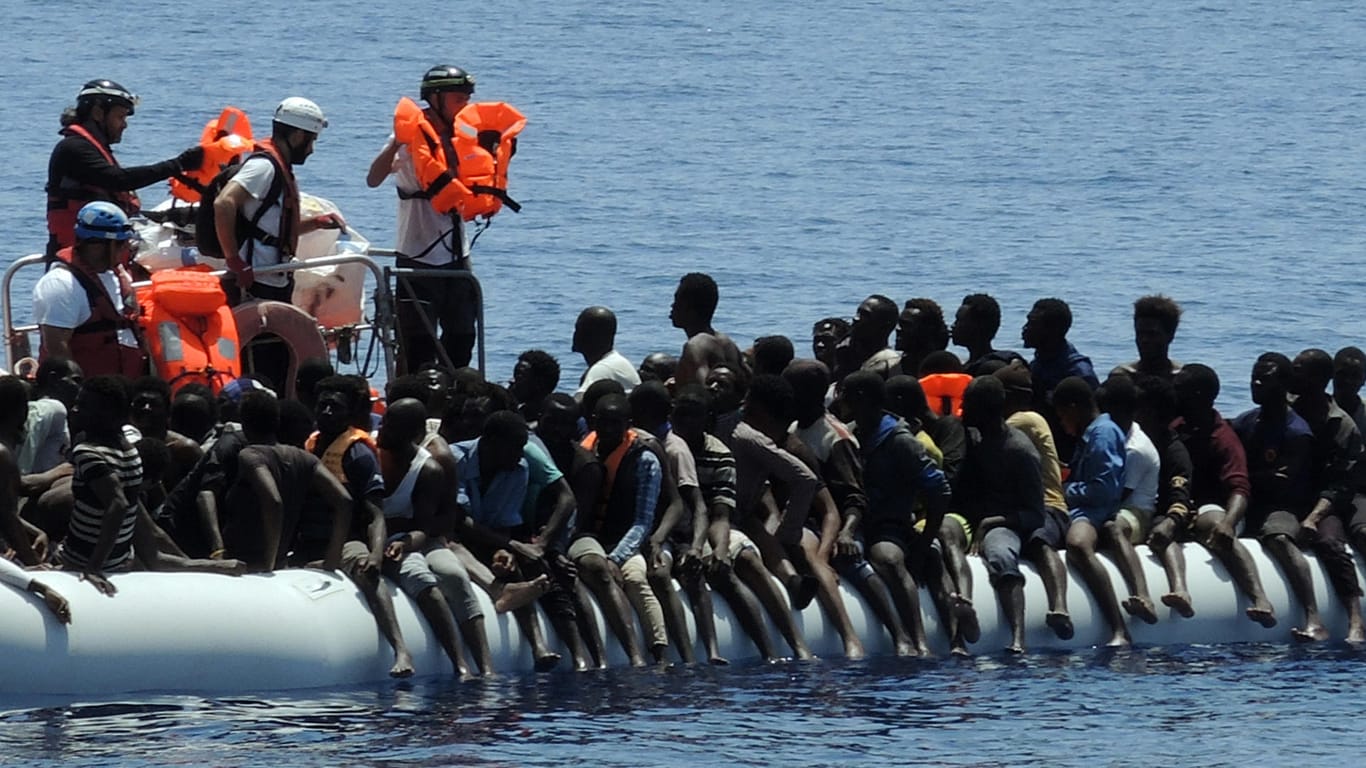 Migranten sitzen in einem überfüllten Schlauchboot auf dem Mittelmeer. Helfer der Hilfsorganisationen Ärzte ohne Grenzen und SOS Méditerranée nähern sich ihnen und verteilen Schwimmwesten.