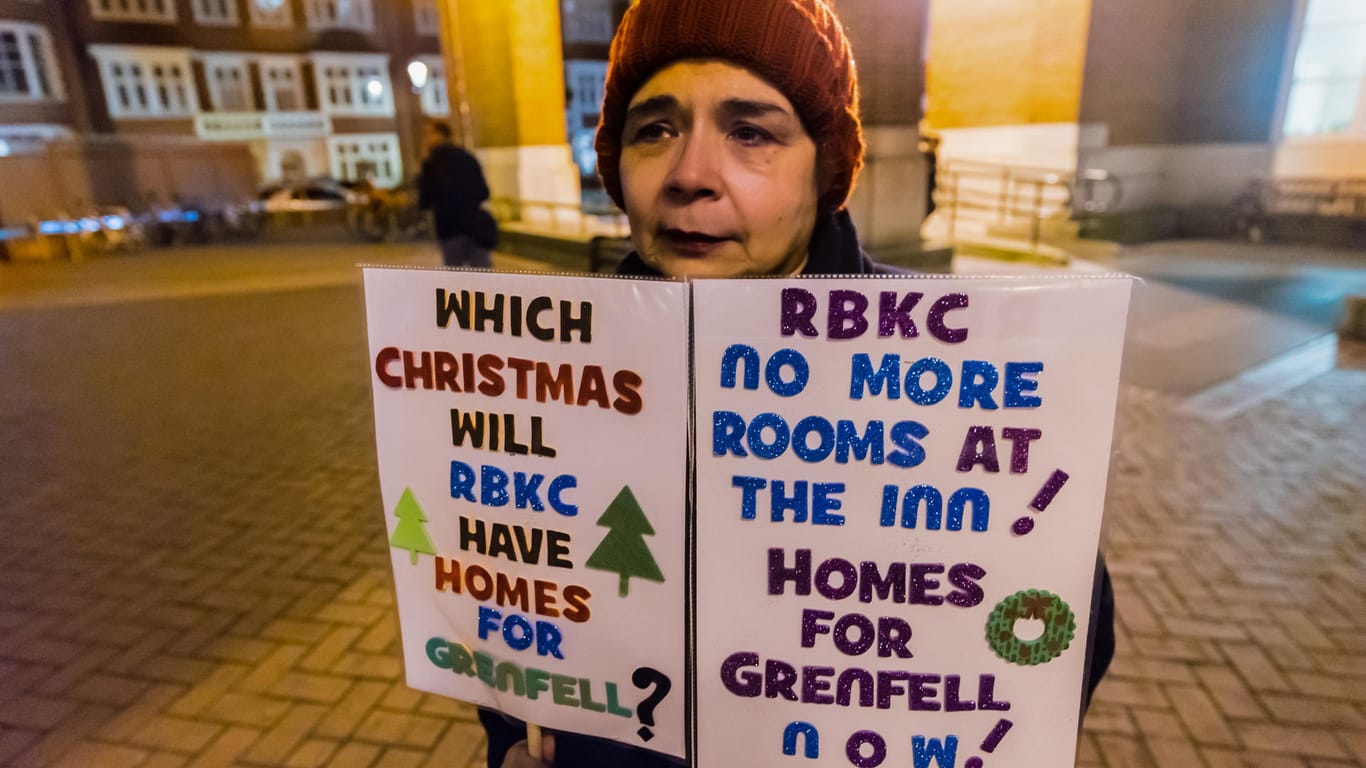 Eine Frau, die Plakate mit der Aufschrift "Which Christmas will have homes for Grenfell? ... Homes For Grenfell now" bei einem Protest zeigt: Bei dem verheerenden Brand im Grenfell-Hochhaus starben vor einem halben Jahr 71 Menschen.