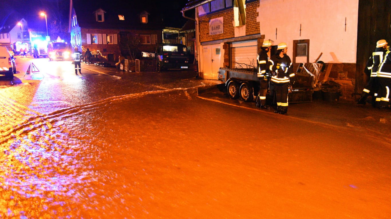 Einsatzkräfte der Feuerwehr stehen auf einer überfluteten Straße. Ein Bach ist über die Ufer getreten und hat die Ortsdurchfahrt geflutet.