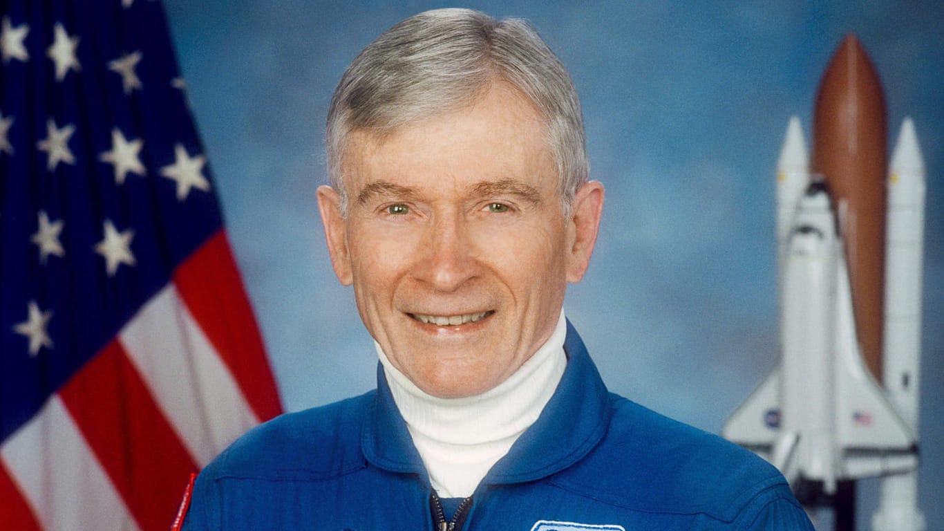 John Young posiert in NASA-Montur vor einem Space Shuttle-Modell.