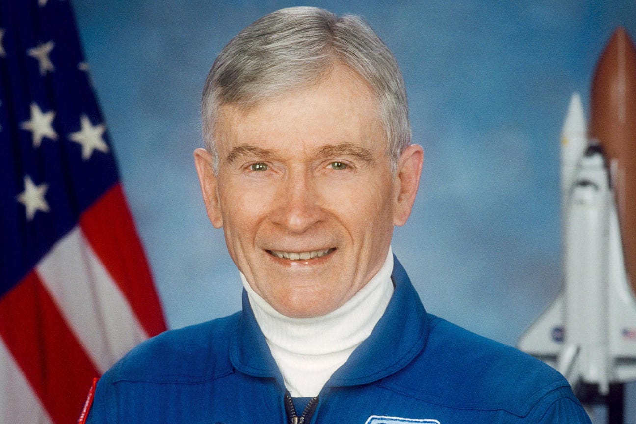 John Young posiert in NASA-Montur vor einem Space Shuttle-Modell.