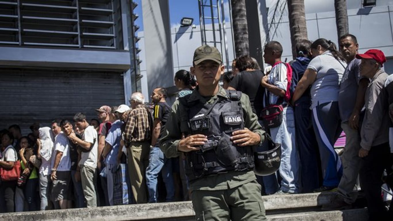 Zahlreiche Menschen stehen in Caracas stundenlang Schlange vor einem Lebensmittelladen, der von einem Polizisten bewacht wird.