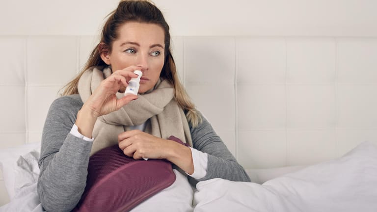 Kranke Frau mit Nasenspray im Bett: Die Anwendung von Nasenspray bei einer Erkältung ist mittlerweile zur Normalität geworden. Allerdings auch die darauf folgende Sucht.