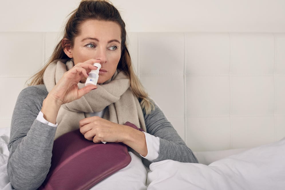 Kranke Frau mit Nasenspray im Bett: Die Anwendung von Nasenspray bei einer Erkältung ist mittlerweile zur Normalität geworden. Allerdings auch die darauf folgende Sucht.