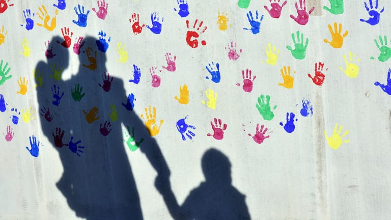 Schatten von Mann mit zwei Kindern auf Wand mit bunten Handabdrücken: Dem "Kinderreport 2018" zufolge werden Familien mit höheren Einkommen bislang stärker entlastet als Familien mit niedrigen Einkommen.
