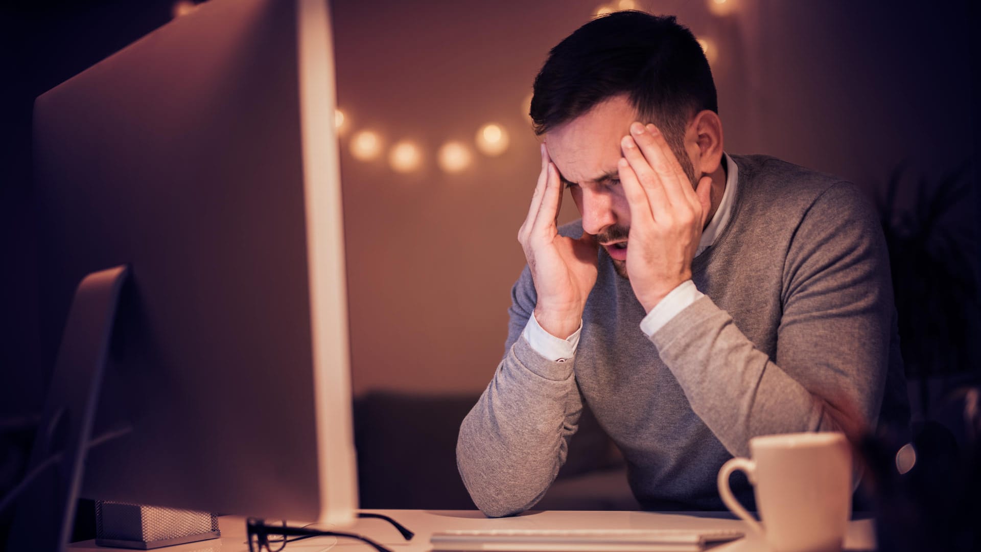 Mann sitzt mit Händen an den Schläfen und angestrengtem Gesicht am Laptop im Halbdunkel