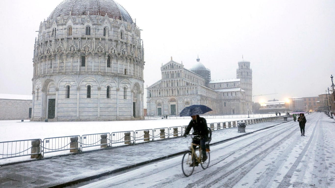 Italien, Pisa: Ein Fahrradfahrer fährt im Schneegestöber mit Schirm eine Straße entlang. Im Hintergrund steht der "Schiefe Turm von Pisa".