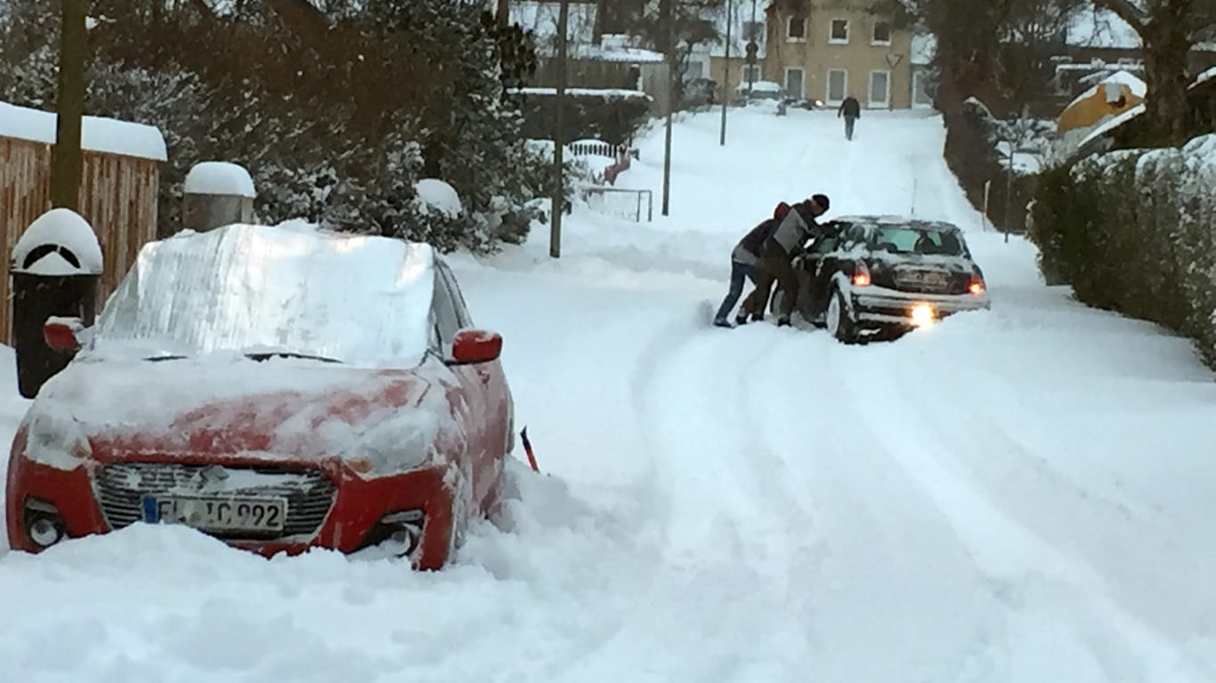 Passanten in Flensburg schieben ein Auto an, das sich auf einer schneebedeckten Straße festgefahren hatte. Besonders im Großraum Flensburg sorgten Schneeverwehungen von zum Teil über einem Meter dafür, dass Straßen nicht befahrbar waren.