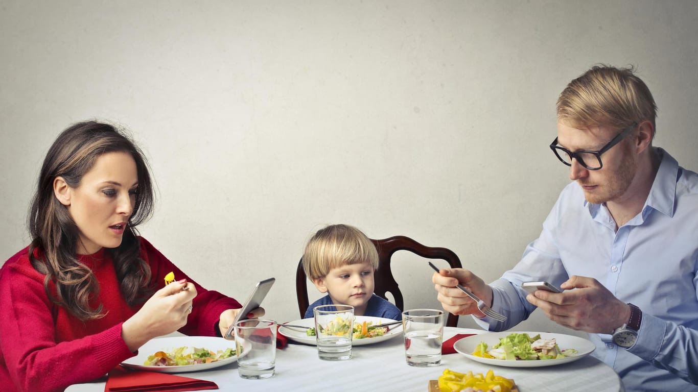 Eltern mit Kleinkind beim Essen. Beide halten ein Mobiltelefon in der Hand.
