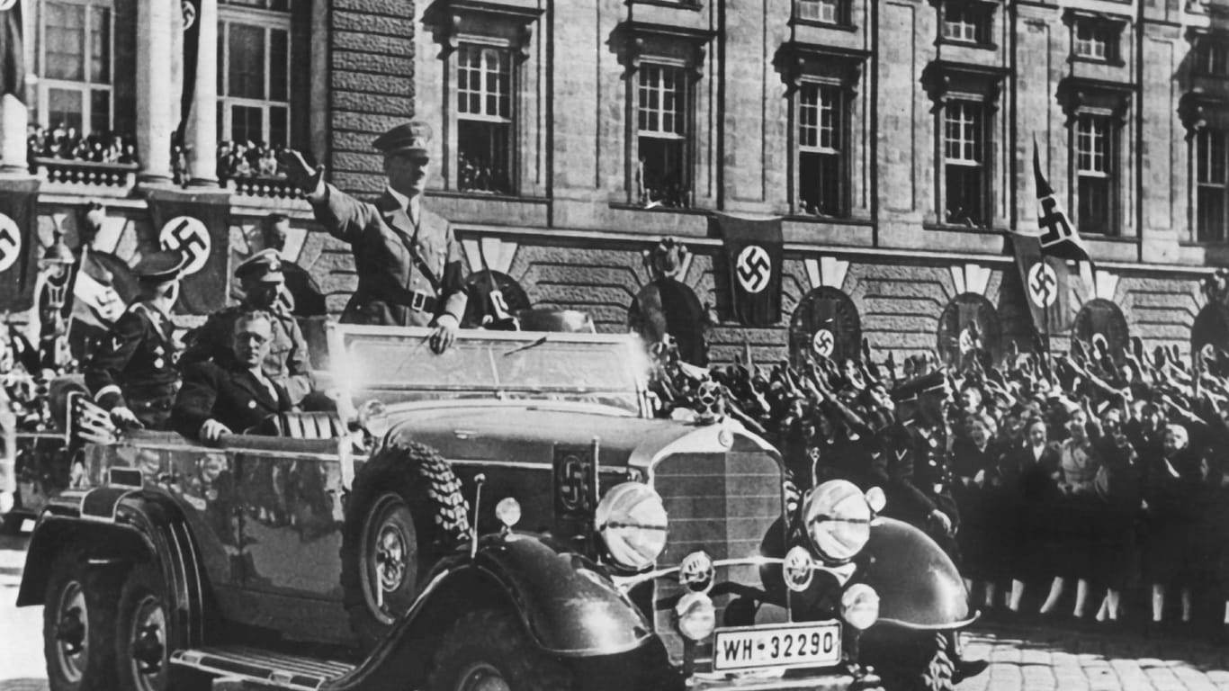 In einem Auto stehend grüßt Adolf Hitler die ihm zujubelnde Menschenmenge in Wien (Österreich) am 14. März 1938, neben ihm sitzt der österreichische Kanzler Arthur Seyss-Inquart: Am 12. März 1938 überschreiten Soldaten der deutschen Wehrmnacht die Grenze zur Alpenrepublik.