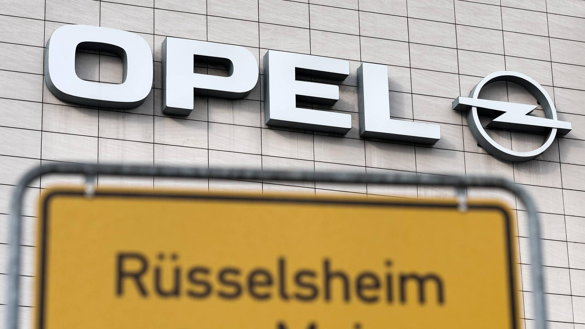 Das Straßenschild von Rüsselsheim am Main vor dem Opel-Logo. Die Stadt ist untrennbar mit dem Autobauer verbunden.