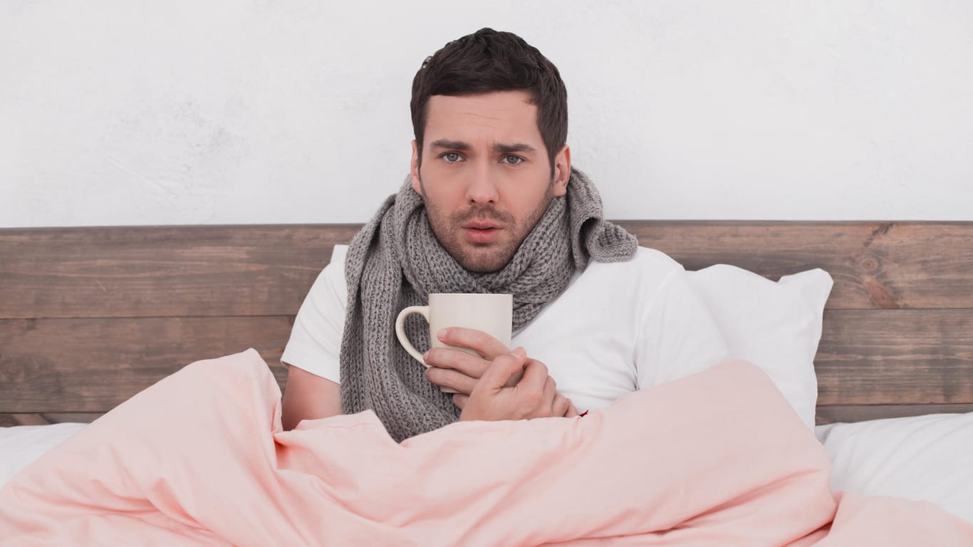 Mann um die 30 sitzt mit Schal und Tee im Bett.