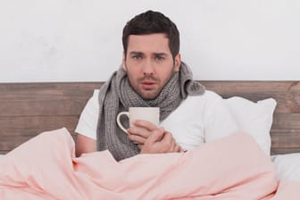 Mann um die 30 sitzt mit Schal und Tee im Bett.