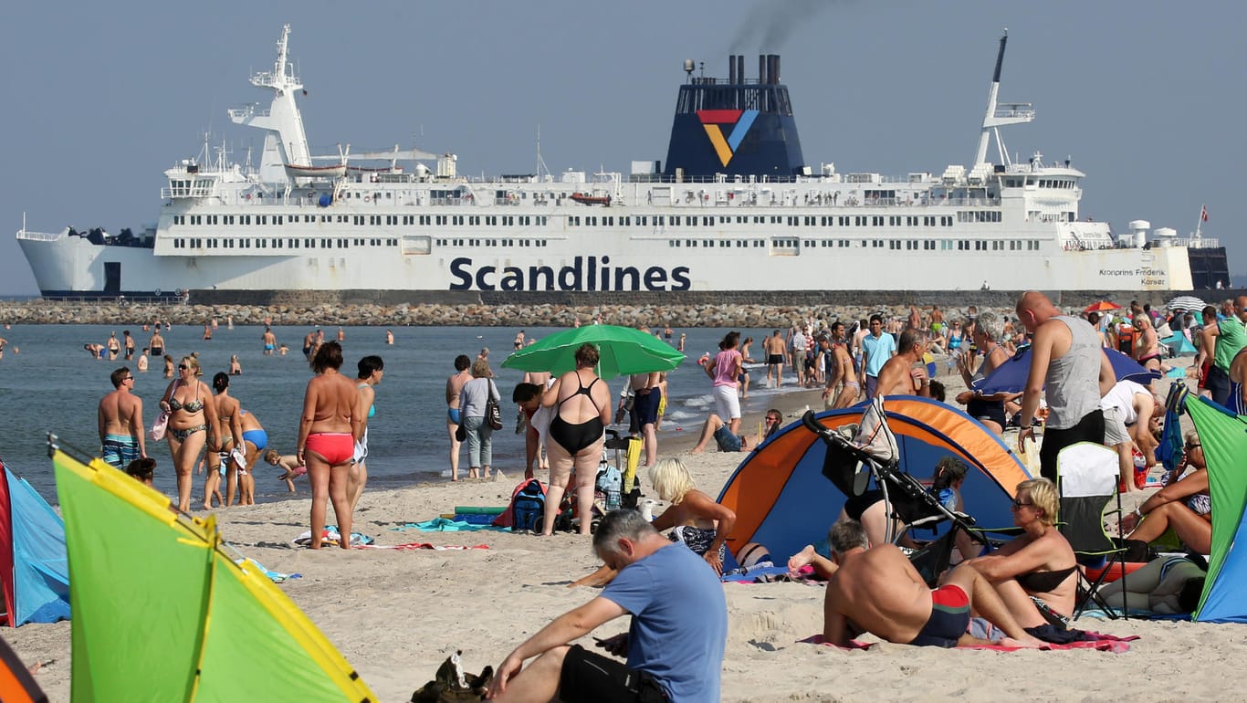 Badegäste sitzen am Ostseestrand von Warnemünde (Mecklenburg-Vorpommern) und im Hintergrund läuft eine Scandlines-Fähre nach Gedser (Dänemark) aus: Deutsch-dänische Fährreederei für 1,7 Milliarden Euro verkauft.