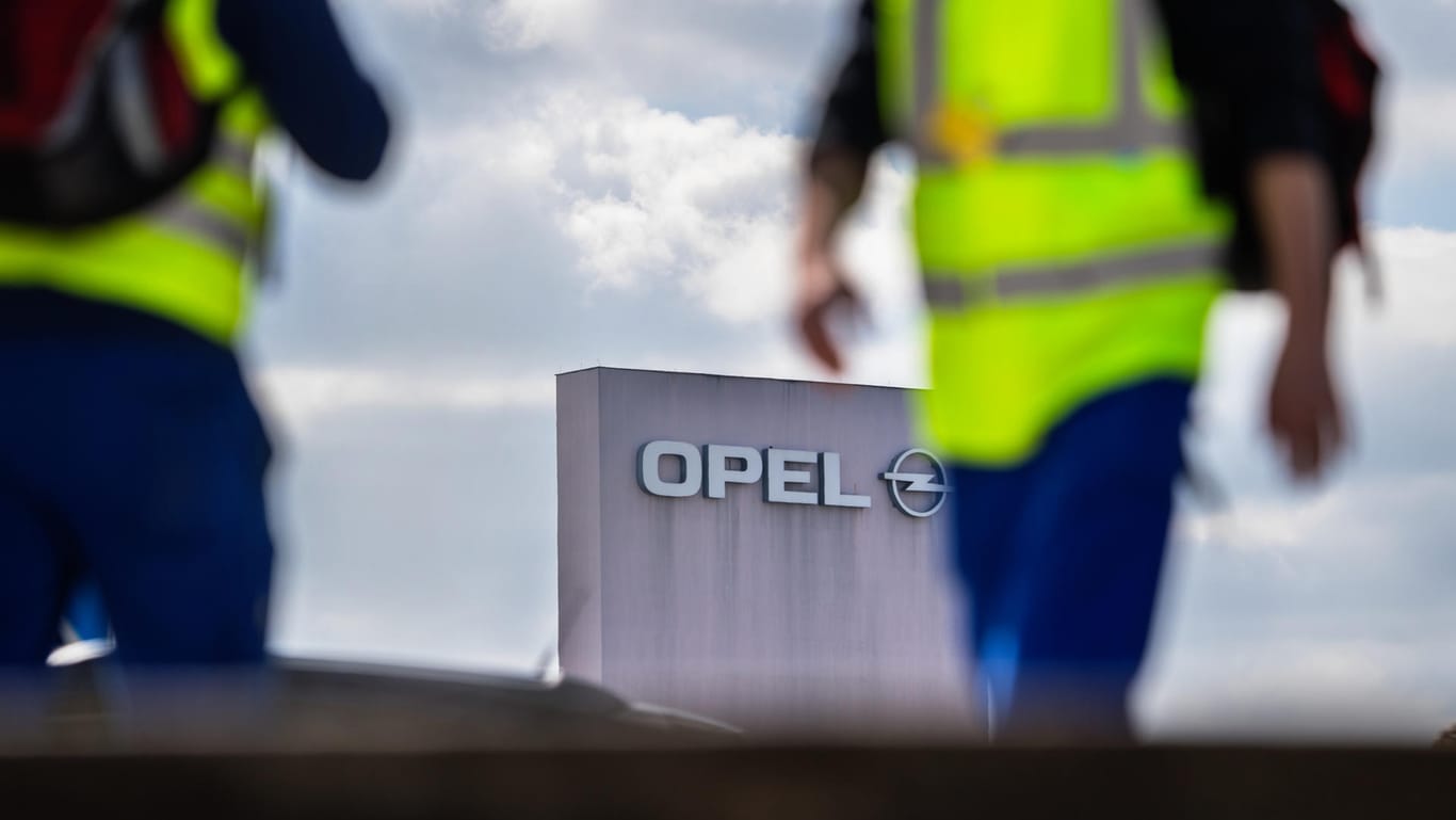 Zwei Männer in Sicherheitswesten gehen in Rüsselsheim (Hessen) am Stammwerk von Opel vorbei: Opel legt freiwilliges Abfindungsprogramm für Mitarbeiter auf.