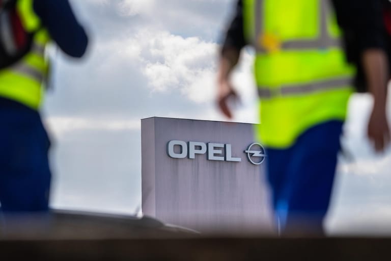 Zwei Männer in Sicherheitswesten gehen in Rüsselsheim (Hessen) am Stammwerk von Opel vorbei: Opel legt freiwilliges Abfindungsprogramm für Mitarbeiter auf.