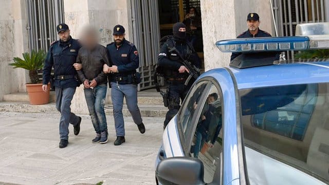 Einer der Verdächtigen im Falle der Verhaftung fünf mutmaßlicher Komplizen von Anis Amri wird von zwei Polizisten aus der Polizeizentrale in Latina eskortiert. Einer der Verdächtigen im Falle der Verhaftung fünf mutmaßlicher Komplizen von Anis Amri wird von zwei Polizisten aus der Polizeizentrale in Latina eskortiert.