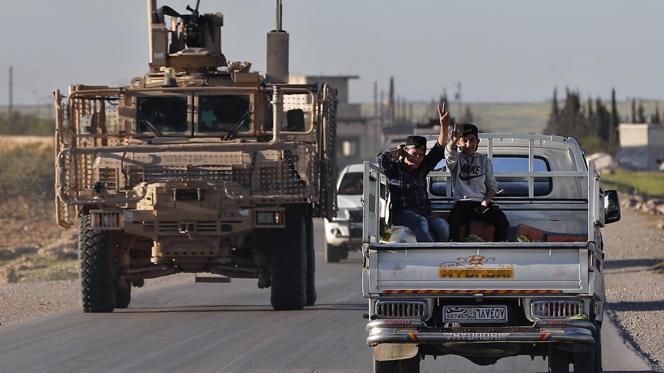 Syrische Jungen auf einem Kleintransporter in Manbidsch neben einem Fahrzeug der US-Truppen: Die USA werden vorerst weiter im Bürgerkriegsland bleiben.