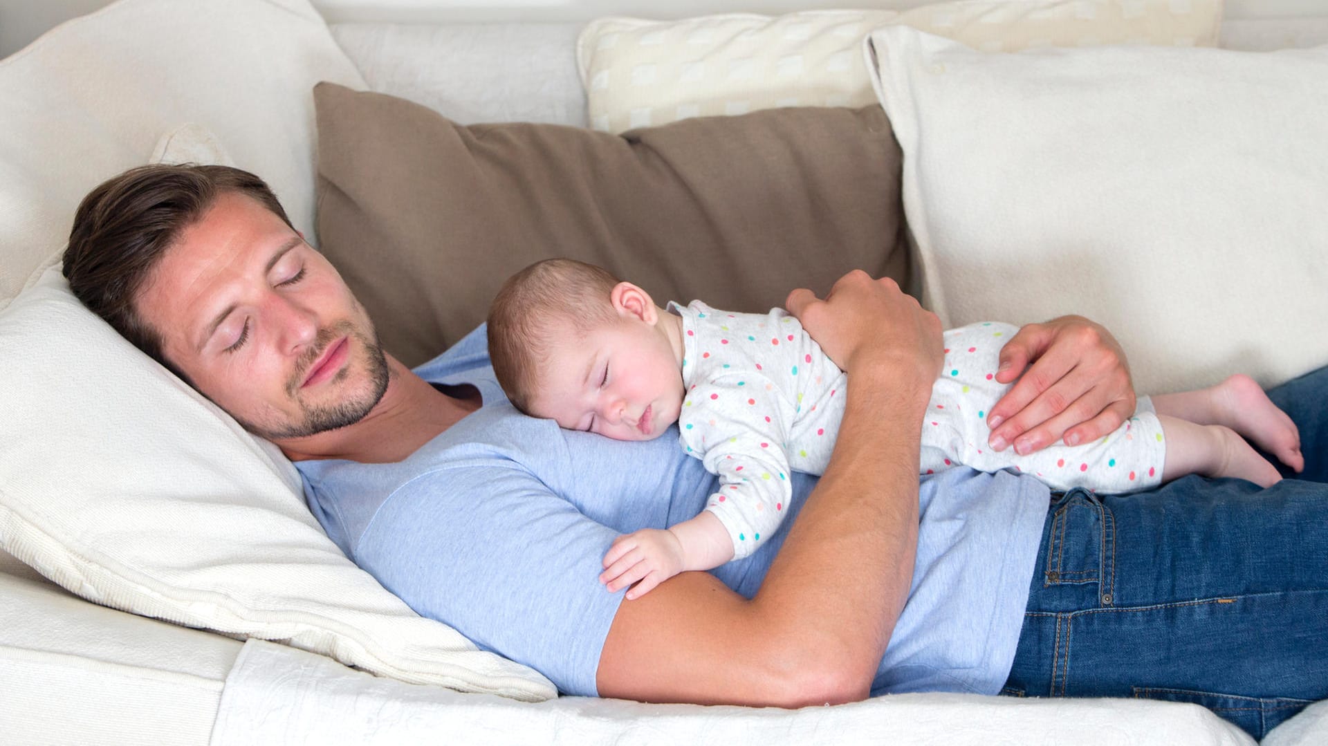 Ein Vater liegt mit seinem Baby auf dem Bauch auf einem Sofa: Sicherer schläft ein Säugling im eigenen Bett.