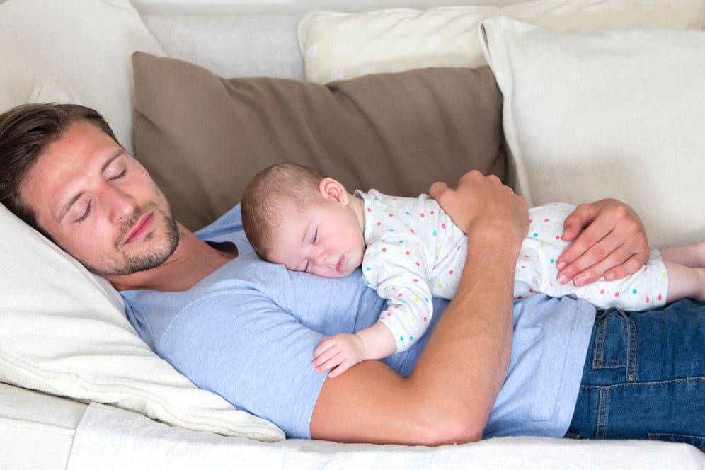 Ein Vater liegt mit seinem Baby auf dem Bauch auf einem Sofa: Sicherer schläft ein Säugling im eigenen Bett.