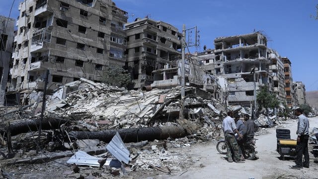 Menschen stehen vor zerstörten Gebäuden in Duma. Menschen stehen vor zerstörten Gebäuden in Duma.