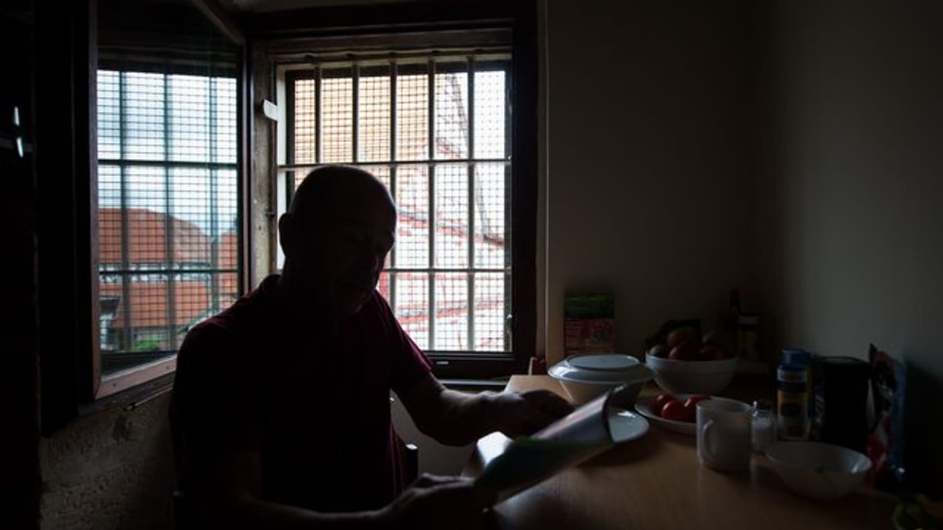 Ein älterer Inhaftierter sitzt vor einem Gitterfenster in seiner Zelle und liest eine Broschüre.