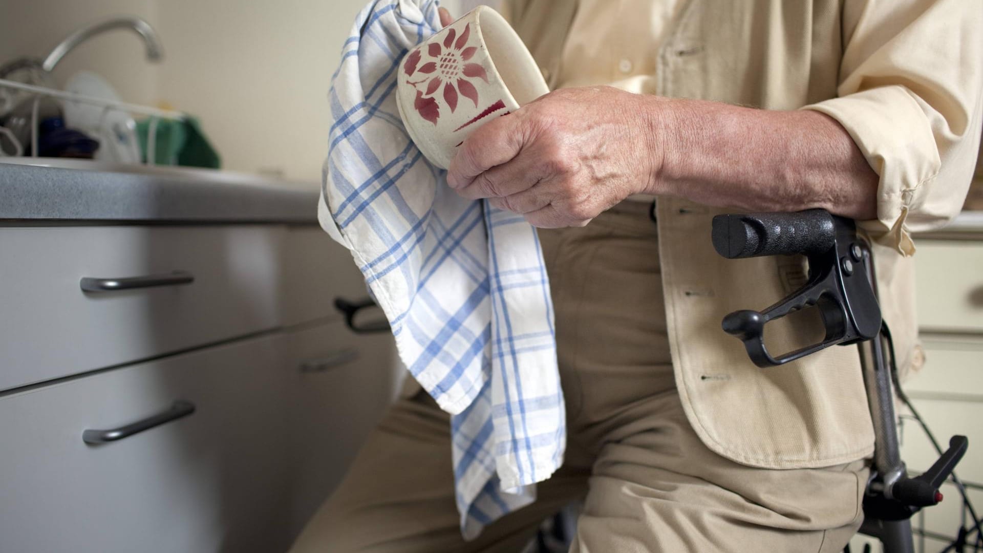 Ein Mann mit Rollator trocknet Geschirr in seiner Küche ab: Wer Unterstützung im Alltag braucht, kann das Finanzamt an den Kosten beteiligen.