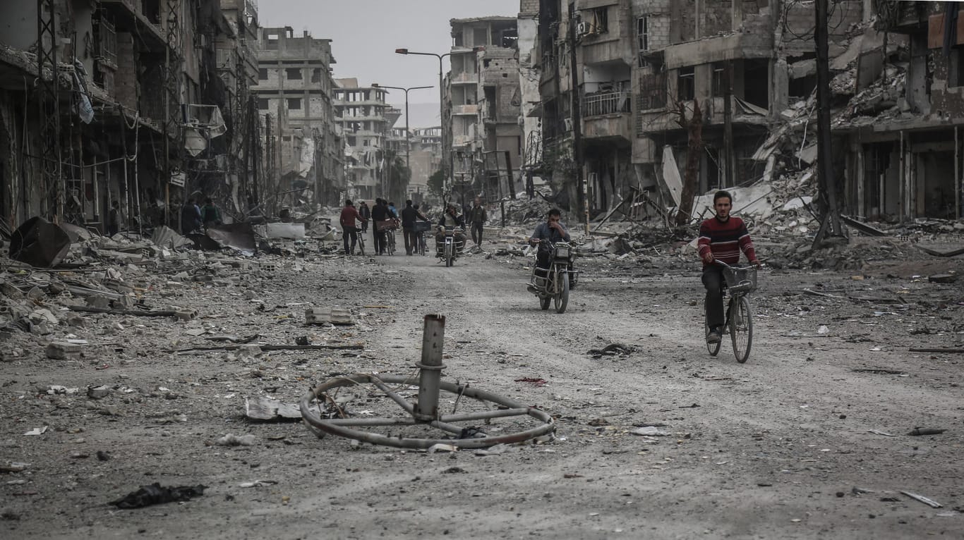 Menschen auf Fahr- und Motorrädern fahren durch eine zerstörte Straße in Ost-Ghuta: In Syrien kämpft der Iran an der Seite vom Machthaber Assad, Russland und der libanesischen Hisbollah.