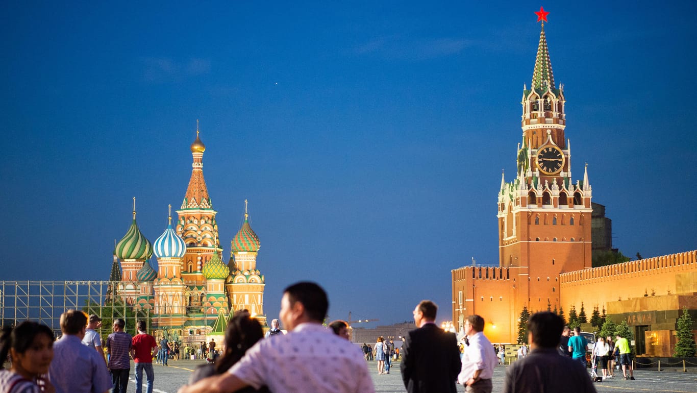 Moskau in Russland: Besucher spazieren zur blauen Stunde auf dem Roten Platz. Links steht die Basilius-Kathedrale und rechts das Lenin-Mausoleum und der Moskauer Kreml.