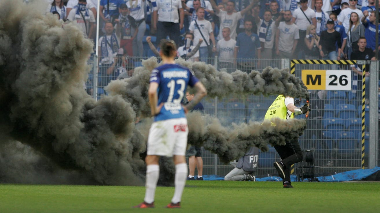 Unfassbare Szene: Ein Ordner räumt qualmende Pyrotechnik vom Feld, ein Spieler schaut fassungslos zu.