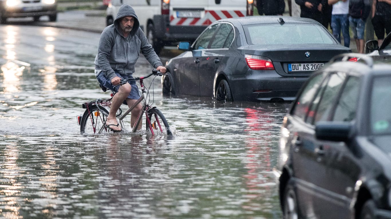 Duisburg: Ein Radfahrer fährt über eine überflutete Straße an liegengebliebenen Autos vorbei: Heftige Unwetter ziehen über Nordrhein-Westfalen.