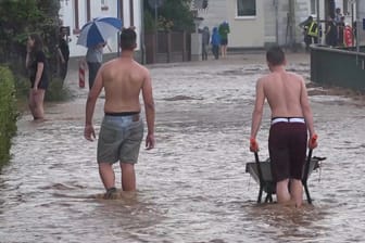 Hessen, Etzen-Gesäß: Männer waten mit einer Schubkarre durch eine überflutete Straße: Die kleine Ortschaft im Odenwaldkreis ist am Donnerstagabend nach einem schweren Unwetter überflutet worden.