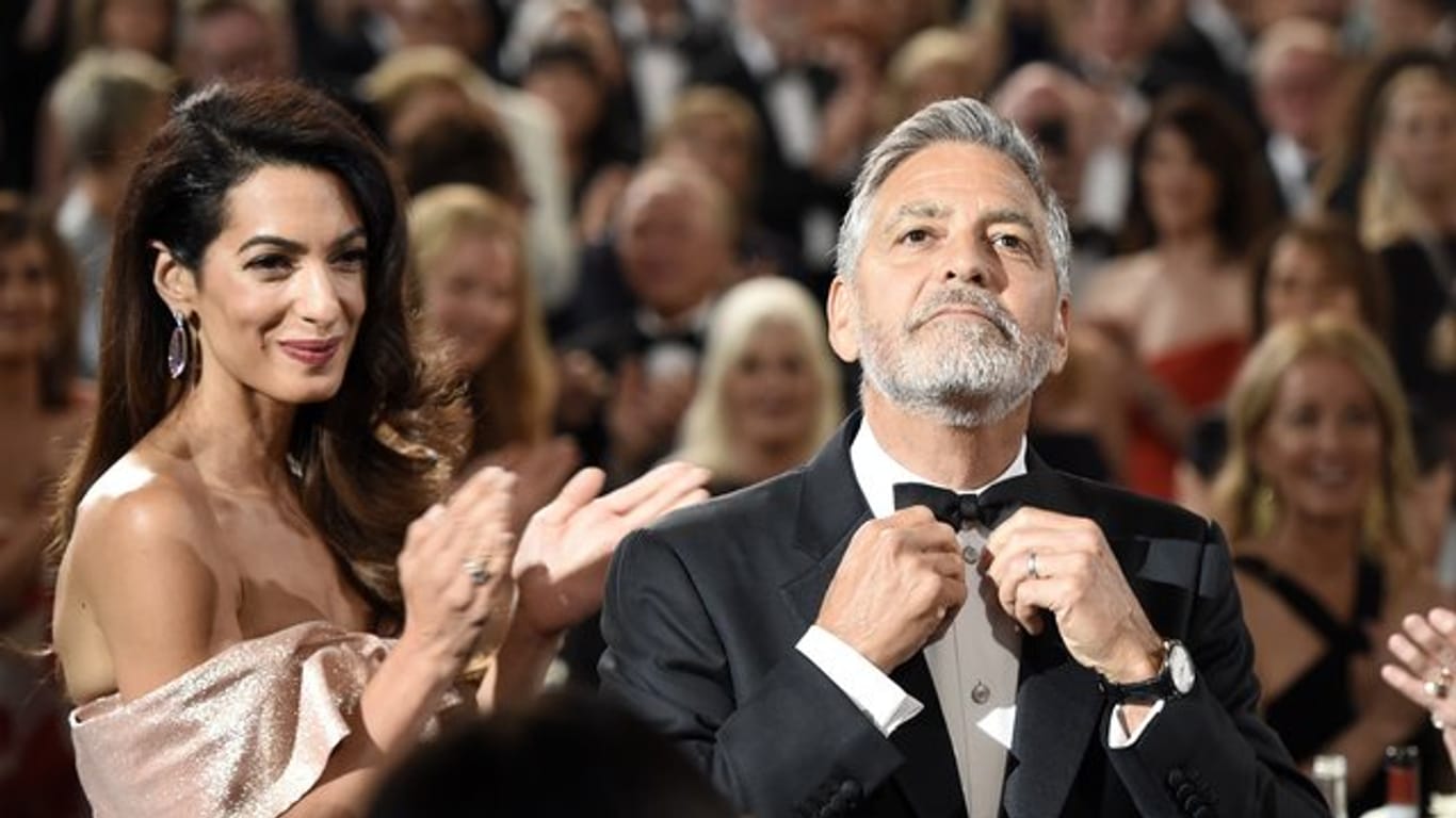 George Clooney richtet seine Fliege, seine Frau Amal applaudiert.